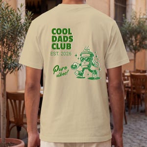 Pode incluir: Camiseta bege com um gráfico verde que diz "COOL DADS CLUB EST. 2026" e "Pure vibes!". Também apresenta uma xícara de café de desenho animado usando óculos de sol e dando um joinha. É uma roupa casual.