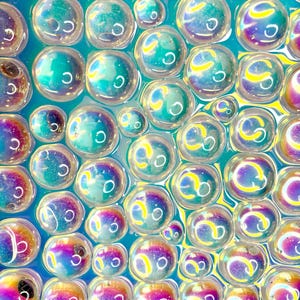 以下が含まれることがあります： 密集したパターンで配置された、虹色に輝く球状の泡の集合体。それぞれの泡は、青、紫、黄色の色合いが変化する半透明の表面を持ち、表面全体に明るい円形のハイライトが光を反射しています。