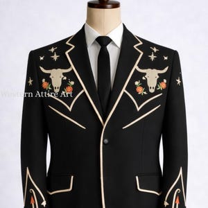 Könnte beinhalten: Schwarzer Blazer mit cremefarbenen Zierleisten und gestickten Details. Der Blazer hat Stierschädel-, Blumen- und Sternmotive. Die Ärmel haben Pfeildesigns. Ein weißes Hemd und eine schwarze Krawatte werden darunter getragen. Der Blazer wird auf einer Schaufensterpuppe präsentiert.