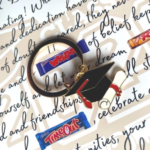 May include: A gold-toned metal keychain featuring a black graduation cap with a red tassel and a rolled diploma tied with a red ribbon. The keychain is positioned over a background with printed text and several small chocolate candy bars.