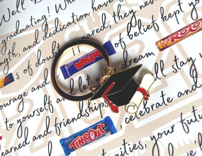 May include: A gold-toned metal keychain featuring a black graduation cap with a red tassel and a rolled diploma tied with a red ribbon. The keychain is positioned over a background with printed text and several small chocolate candy bars.