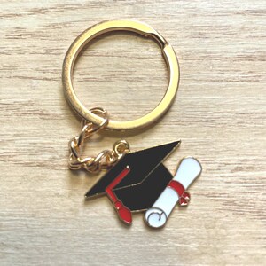 May include: A gold-toned metal keychain featuring a charm shaped like a black graduation cap with a red tassel and a white diploma tied with a red ribbon. The charm is attached to a circular gold-toned split ring by a short metal chain link.