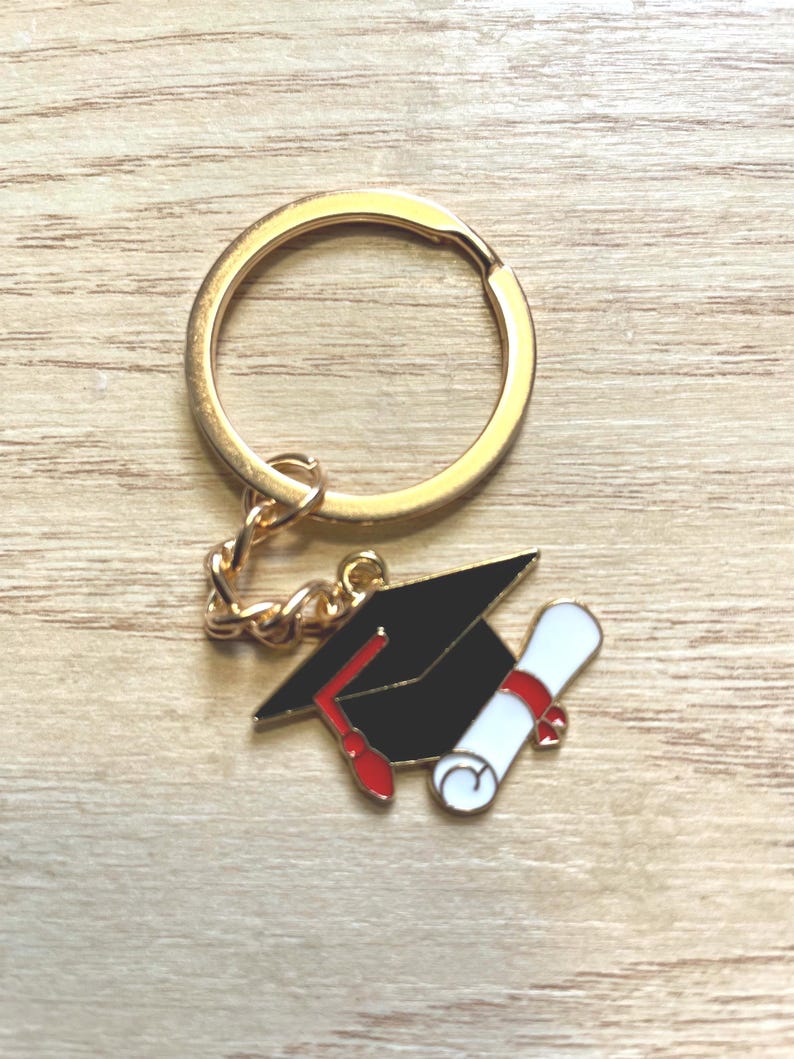 May include: A gold-toned metal keychain featuring a charm shaped like a black graduation cap with a red tassel and a white diploma tied with a red ribbon. The charm is attached to a circular gold-toned split ring by a short metal chain link.