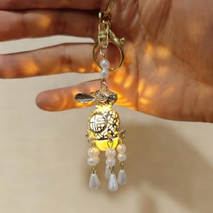 Puede incluir: Un llavero de metal dorado con un diseño de conejo y un interior brillante. El conejo tiene un patrón decorativo y está adornado con cuentas de perlas blancas. El llavero incluye un cierre y una pequeña cadena con un acento de perla.