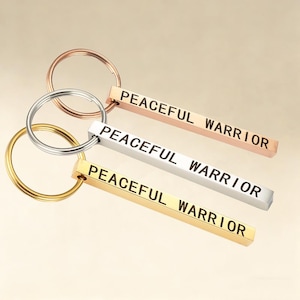 Peut inclure: Trois porte-clés avec l'inscription "PEACEFUL WARRIOR" gravée sur des barres rectangulaires. Les porte-clés sont de couleur or, argent et or rose, chacun attaché à un anneau porte-clés assorti. Le fond est de ton neutre.