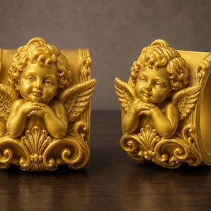 May include: Two gold-colored napkin rings, each featuring a sculpted cherub with curly hair and wings. The cherubs are positioned with hands clasped under their chins, set against a decorative, ornate base with scrollwork details.