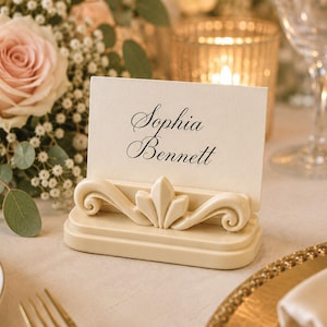 May include: A cream-colored rectangular place card holder with a decorative scroll and leaf-like center design. A white rectangular card is inserted into the holder, featuring the printed black text Sophia Bennett in a classic serif script font.