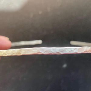 May include: A silver-colored, hammered metal bracelet. The bracelet has a textured surface and is open at both ends. The metal has a slight gradient of color, transitioning from a golden hue to a silver tone. The bracelet is shown against a dark background.