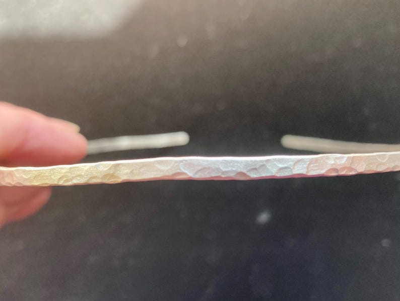 May include: A silver-colored, hammered metal bracelet. The bracelet has a textured surface and is open at both ends. The metal has a slight gradient of color, transitioning from a golden hue to a silver tone. The bracelet is shown against a dark background.