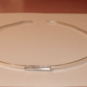 May include: A silver-colored choker necklace. The necklace is a simple, thin band with a rectangular clasp in the center. The necklace is displayed on a white surface.