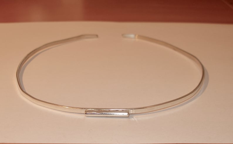 May include: A silver-colored choker necklace. The necklace is a simple, thin band with a rectangular clasp in the center. The necklace is displayed on a white surface.