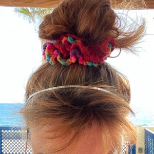 May include: A red, multi-colored crochet scrunchie sits atop a person's head. The scrunchie features a mix of red, orange, purple, and teal yarn. A silver headband is visible below the scrunchie. The background shows a blue ocean and sky.