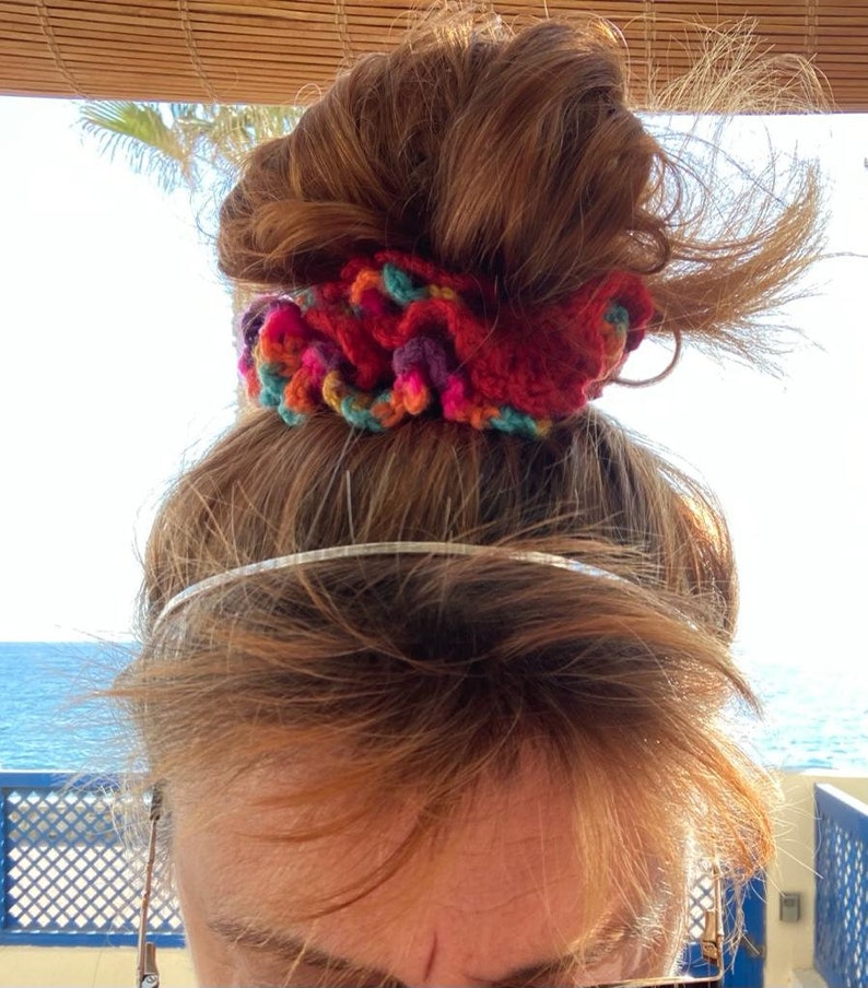May include: A red, multi-colored crochet scrunchie sits atop a person's head. The scrunchie features a mix of red, orange, purple, and teal yarn. A silver headband is visible below the scrunchie. The background shows a blue ocean and sky.