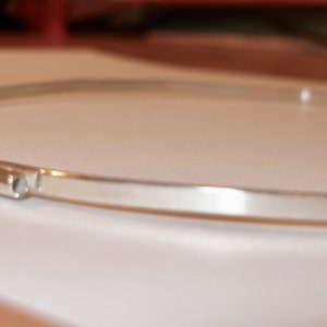 May include: A minimalist silver-colored choker necklace. The necklace has a simple, sleek design with a thin, curved band and a small, cylindrical clasp. The jewelry is displayed on a white surface.