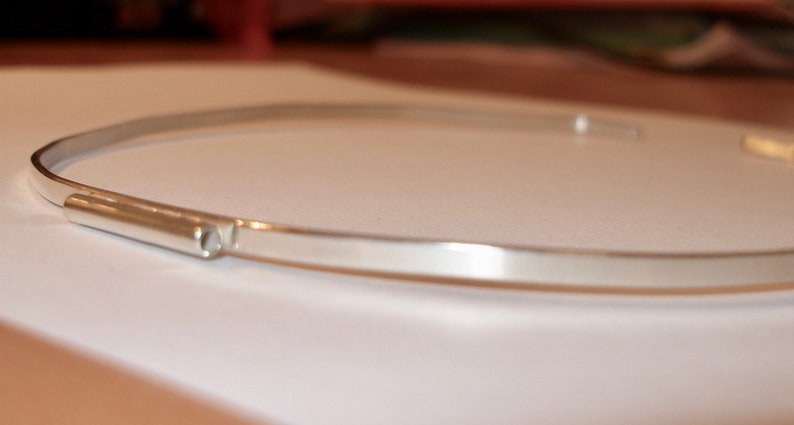 May include: A minimalist silver-colored choker necklace. The necklace has a simple, sleek design with a thin, curved band and a small, cylindrical clasp. The jewelry is displayed on a white surface.