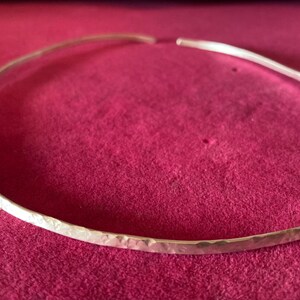 May include: A hammered silver choker necklace. The open-ended collar is displayed on a deep pink surface. The necklace has a textured, hammered finish, reflecting light. This minimalist piece is suitable for everyday wear or special occasions.