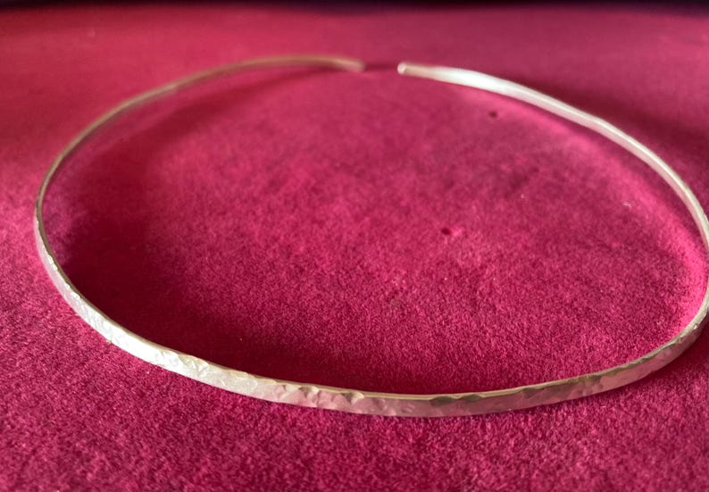 May include: A hammered silver choker necklace. The open-ended collar is displayed on a deep pink surface. The necklace has a textured, hammered finish, reflecting light. This minimalist piece is suitable for everyday wear or special occasions.