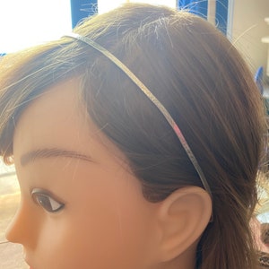 May include: A thin, silver headband worn on a mannequin with brown hair. The headband is a simple, elegant accessory, likely made of metal. The mannequin's skin is a light tone, and the background is blurred.