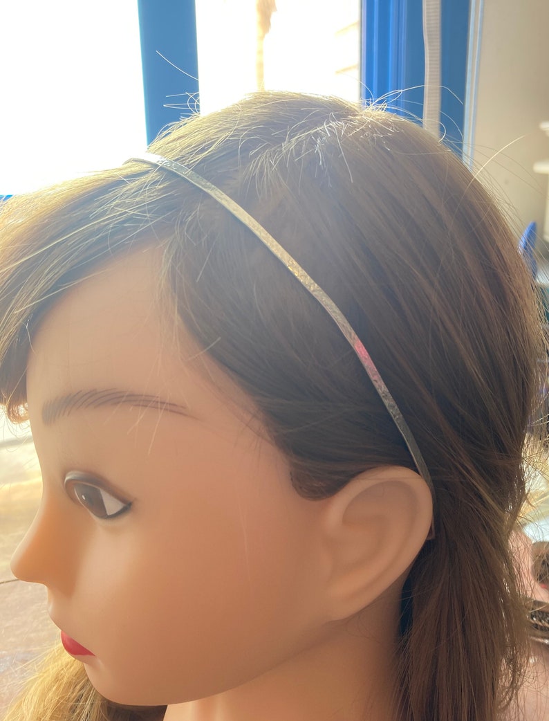 May include: A thin, silver headband worn on a mannequin with brown hair. The headband is a simple, elegant accessory, likely made of metal. The mannequin's skin is a light tone, and the background is blurred.
