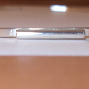 May include: A silver-colored, minimalist choker necklace. The necklace features a thin, flat band with a rectangular bar at the center. The jewelry is displayed on a white surface, with a blurred brown background.