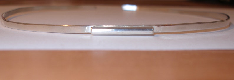 May include: A silver-colored, minimalist choker necklace. The necklace features a thin, flat band with a rectangular bar at the center. The jewelry is displayed on a white surface, with a blurred brown background.