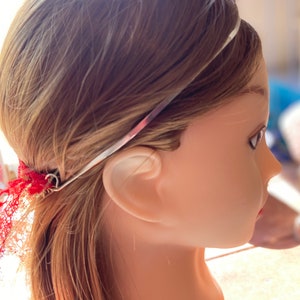 May include: A silver headband with a red and pink floral design is worn on a mannequin head with light brown hair. Red lace is attached to the headband near the ear.