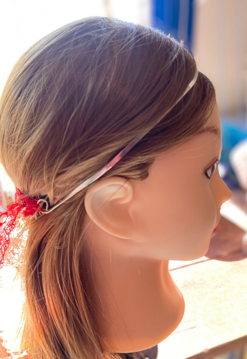 May include: A silver headband with a red and pink floral design is worn on a mannequin head with light brown hair. Red lace is attached to the headband near the ear.