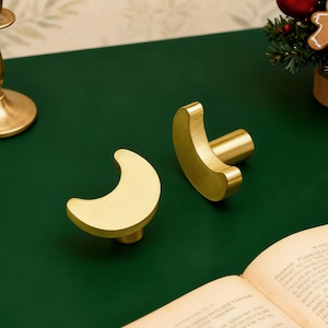 Może przedstawiać: Dwa mosiężne uchwyty do szafek w kształcie półksiężyca na ciemnozielonej powierzchni. Zapalona świeca w mosiężnym świeczniku i mała choinka z ozdobami znajdują się w tle. Otwarta książka i biała orchidea są również obecne.