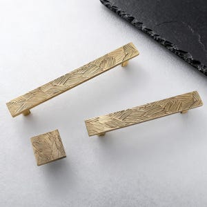 May include: A collection of brass-toned cabinet hardware, including two rectangular pulls and a square knob. Each piece features a textured, cross-hatched pattern. The hardware is displayed on a white surface with a dark, textured accent piece in the background.