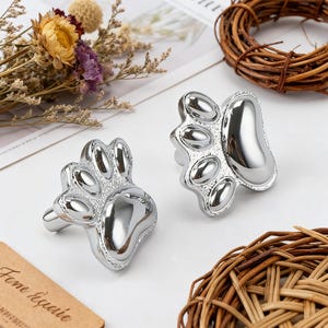 May include: Two shiny silver paw-shaped cabinet knobs. The knobs are highly reflective and detailed, with a paw print design. The knobs are likely for a pet-themed room or furniture. The background includes dried flowers and decorative elements.