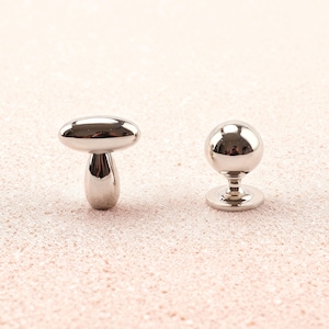 Könnte beinhalten: Zwei glänzende, silberfarbene Metallobjekte. Eines ist ein pilzförmiger Knopf mit abgerundeter Oberseite und zylindrischem Schaft. Das andere ist ein kugelförmiger Knopf mit kreisförmiger Basis. Beide sind reflektierend und liegen auf einer hellrosa Oberfläche.