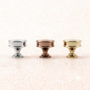 May include: Three decorative cabinet knobs in silver, copper, and gold finishes. Each knob features a rounded top with a circular band and a pedestal base. The knobs are displayed against a light pink background.