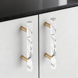 Könnte beinhalten: Zwei Schrankgriffe mit weißem Marmormuster und goldfarbener Hardware. Die zylindrischen Griffe sind an einer weißen Schranktür befestigt. Das Marmormuster weist schwarze Maserungen auf.