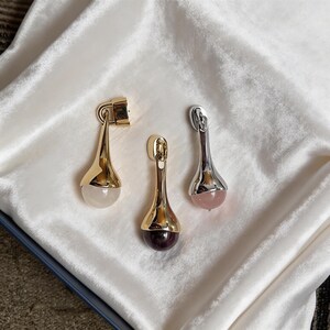 Può includere: Tre orecchini a goccia con finiture in metallo oro e argento. Ogni orecchino presenta una pietra preziosa alla base: un quarzo trasparente, un viola intenso e una pietra rosa. Gli orecchini sono esposti su una superficie di raso bianco.