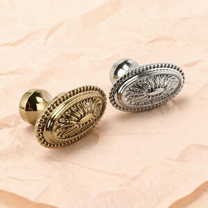May include: Two decorative oval cabinet knobs. One is gold-toned, the other is silver-toned. Both knobs feature a detailed floral design within an oval frame, surrounded by a beaded border. The knobs are set against a light pink background.