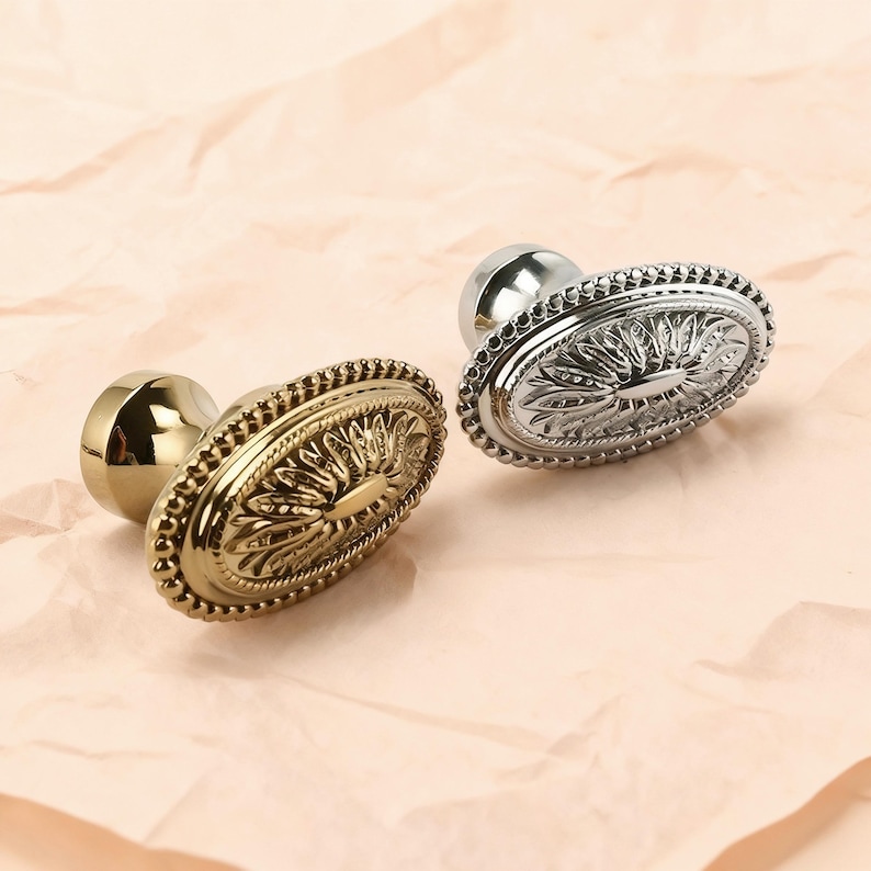 May include: Two decorative oval cabinet knobs. One is gold-toned, the other is silver-toned. Both knobs feature a detailed floral design within an oval frame, surrounded by a beaded border. The knobs are set against a light pink background.