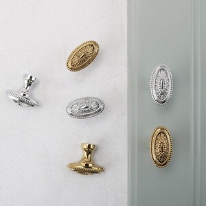 May include: An assortment of decorative cabinet knobs and pulls in gold and silver finishes. The oval-shaped knobs feature a detailed floral design and are displayed on a white and gray background. The knobs are made of metal.