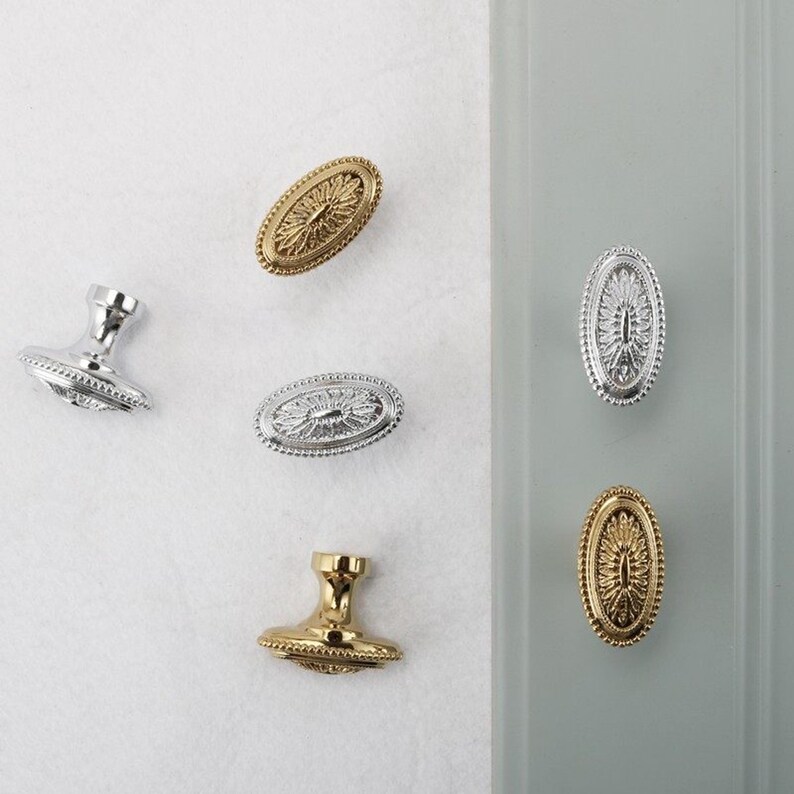 May include: An assortment of decorative cabinet knobs and pulls in gold and silver finishes. The oval-shaped knobs feature a detailed floral design and are displayed on a white and gray background. The knobs are made of metal.