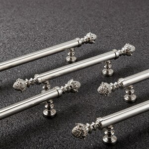 May include: Several silver cabinet pulls with a pineapple-shaped design on each end. The pulls are cylindrical with decorative detailing and are set against a dark, textured background. These hardware pieces are likely for kitchen or bathroom cabinets.