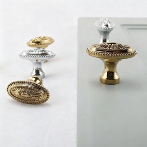 May include: Close-up of several decorative cabinet knobs in gold and silver finishes. The knobs have ornate, detailed designs, including oval and round shapes. The gold knobs have a shiny, polished appearance, while the silver knobs have a more matte finish.