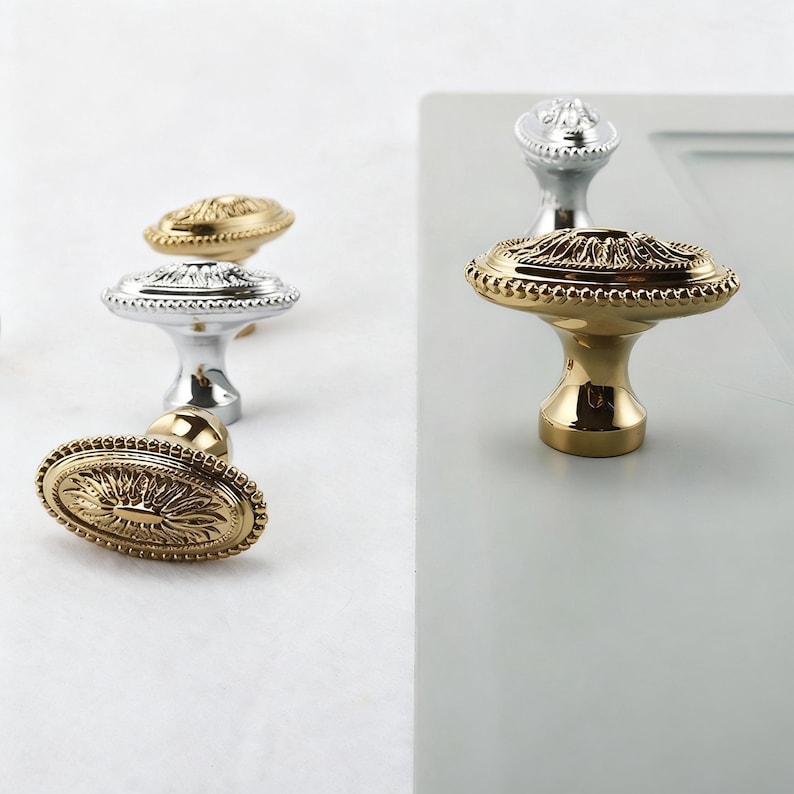 May include: Close-up of several decorative cabinet knobs in gold and silver finishes. The knobs have ornate, detailed designs, including oval and round shapes. The gold knobs have a shiny, polished appearance, while the silver knobs have a more matte finish.