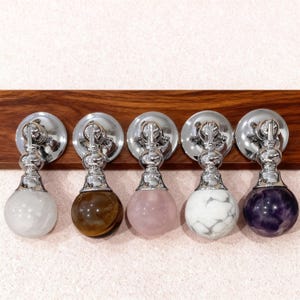 Puede incluir: Cinco tiradores decorativos para armarios con herrajes cromados pulidos y esferas de piedras preciosas. Las esferas son blancas, marrones, rosas, mármol blanco y moradas. Montados en una tabla de madera oscura.
