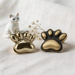 May include: Two decorative paw-shaped cabinet knobs. One is gold-colored, the other is bronze with gold accents. A white ceramic cat figurine and sprigs of white flowers are in the background.