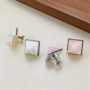 Può includere: Quattro pomelli decorativi per mobili. Due sono bianchi con venature grigie, due sono rosa. I pomelli hanno una forma geometrica a piramide e sono incastonati in basi di metallo dorato o argentato. I pomelli sono su una superficie chiara.