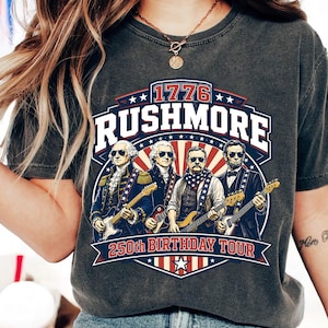 Puede incluir: Una camiseta gris carbón con un estampado gráfico de cuatro figuras históricas tocando guitarras eléctricas. El texto dice 1776, RUSHMORE y 250th BIRTHDAY TOUR. El diseño incorpora colores rojo, blanco y azul con detalles de estrellas.