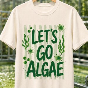 Könnte beinhalten: Ein cremefarbenes Kurzarm-T-Shirt mit einem grünen Grafikdruck und dem Text "LET'S GO ALGAE" in einer fetten, abgenutzten Schriftart. Das Design zeigt Illustrationen von Seetang und mikroskopischen Algenzellen auf hellgrünen vertikalen Streifen.