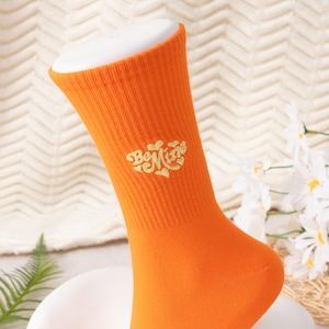 May include: Bright orange crew sock with the embroidered text "Be Mine" in a gold script. The sock is ribbed and has a white form inside. The background is a cream-colored, textured fabric with white flowers.