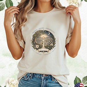 Op de afbeelding: Een beige t-shirt met een cirkelvormige grafische print van een doolhof omringd door bomen, bloemen en een maansikkel. De tekst luidt "PEOPLE • PATHS • POSSIBILITY" en "LISTEN • SUPPORT • EMPOWER • BELONG" met "SMALL STEPS BIG CHANGE" op een banner.