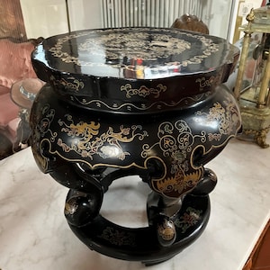 May include: A black, antique, round table with ornate gold detailing. The table has a glossy finish and features intricate designs on the tabletop and sides. The table has a unique, curved base and is likely made of wood.