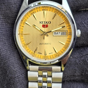 May include: A gold-coloured Seiko automatic wristwatch with a silver stainless steel strap. The watch face has a vertical striped pattern, with the day and date displayed. The watch is marked "SEIKO 5" and "AUTOMATIC".